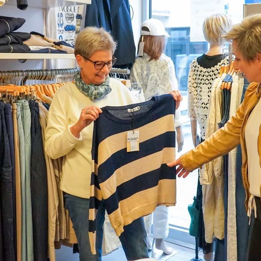 Two women in a clothing store, one holding a striped sweater. Mannequins in the background with clothes on hangers.