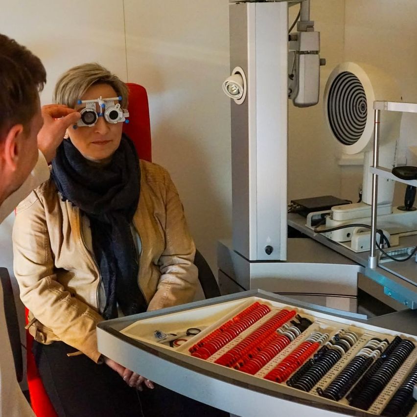 A woman is having her eyes examined by a man wearing a white coat. The woman has a black scarf and wears glasses. The tray contains red and black lenses.