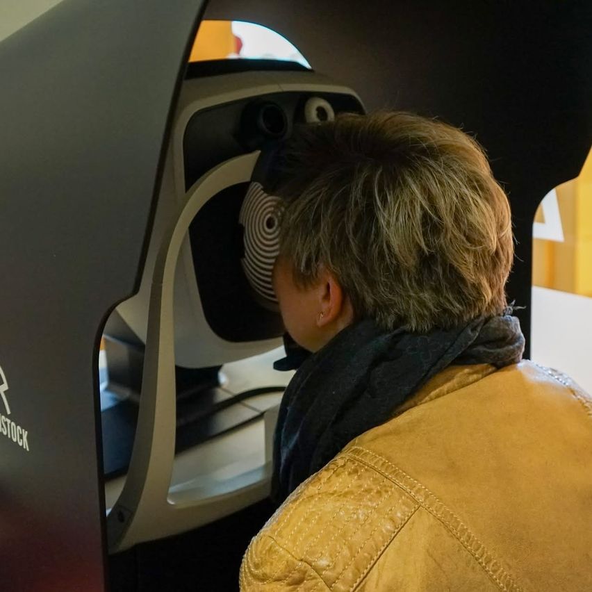 A person uses a facial recognition device, focusing on a camera with a circular pattern. They wear a tan jacket and a scarf.