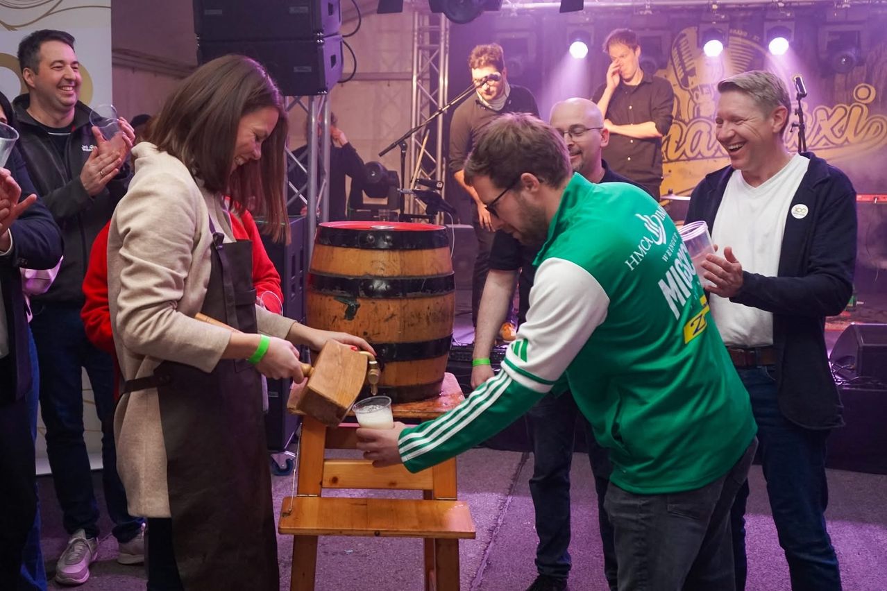 A group of people tap a barrel with a wooden mallet and pour beer into a glass. One woman smiles while a man in a green shirt takes the glass.