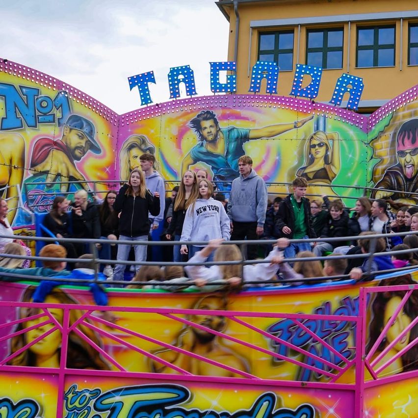 A group of people are watching a colorful ride at an amusement park. The ride has a sign that says 'No.1'. The ride features pictures of people in various poses.