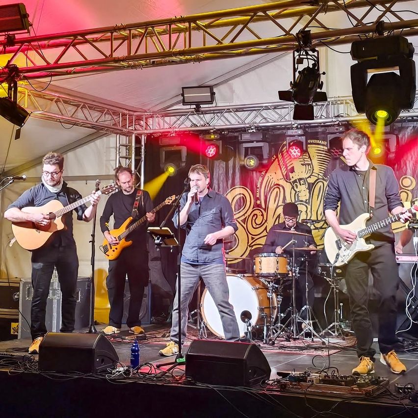 A band performs on a stage under a tent, with a microphone stand, speakers, and musical instruments. The man in the center sings while the others play guitar, drums, and bass.