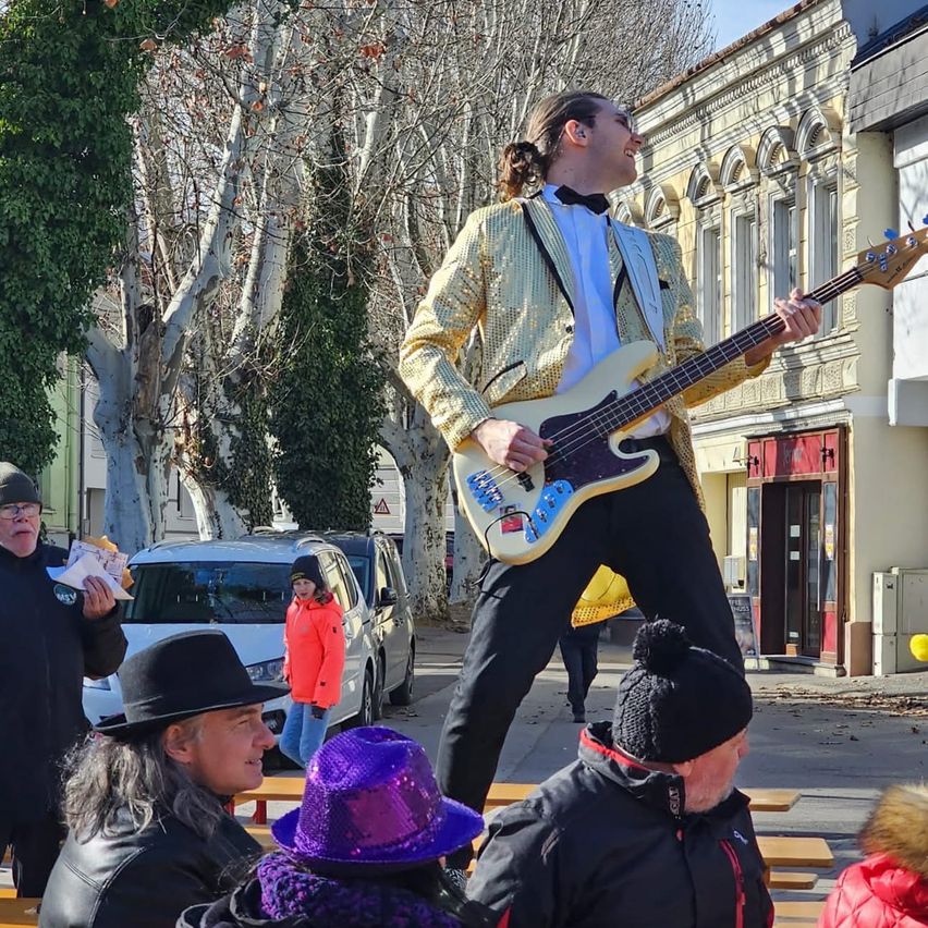 Ein Mann in einem glänzenden Anzug spielt eine Bassgitarre auf einer Straße, während mehrere Menschen zusehen. Hinter ihm ist ein Gebäude mit verzierten Fenstern und einem geparkten Auto zu sehen.
