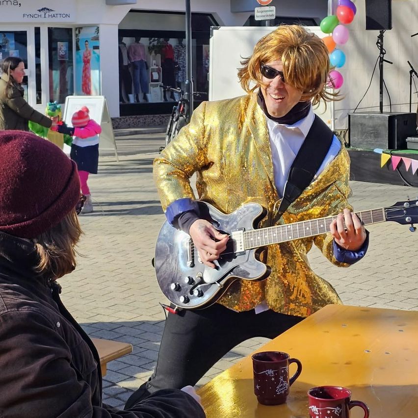 Ein Mann in einem goldenen Anzug spielt Gitarre auf einer Bühne. Eine Frau und ein Kind beobachten. Eine andere Person in einer Mütze sitzt auf einer Bank.