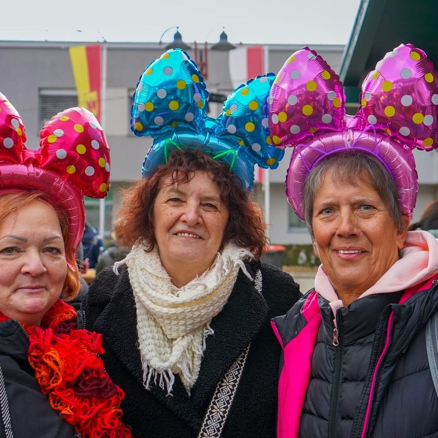 Drei Frauen mit rosa und blauen Minnie Mouse Luftballon-Hüten lächeln für die Kamera. Die Frau in der Mitte hat einen weißen Schal. Hinter ihnen steht ein Gebäude mit Fahnen.
