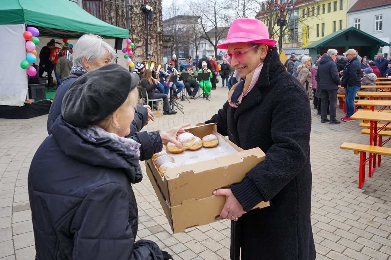 Eine Frau mit einer rosa Hut hält eine Schachtel mit Donuts und gibt sie einer älteren Frau in einer Baskenmütze. Hinter ihnen spielen Musiker, und Menschen versammeln sich.
