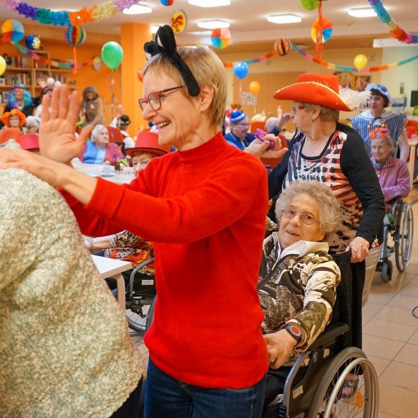 Eine Versammlung älterer Menschen in einem Raum, eine Frau im Rollstuhl winkt einer Frau in einem roten Shirt zu, beide lächeln. Bunte Dekorationen und Ballons schmücken den Raum.