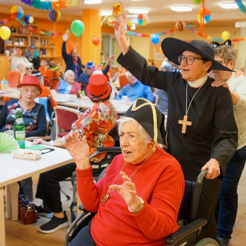 In einer festlichen Umgebung feiern ältere Menschen, einige im Rollstuhl, andere stehend. Eine Frau in einem roten Pullover winkt, während ein Mann in Schwarz hinter ihr ihre Hand hält. Der Raum ist mit bunten Luftballons und Dekorationen geschmückt.
