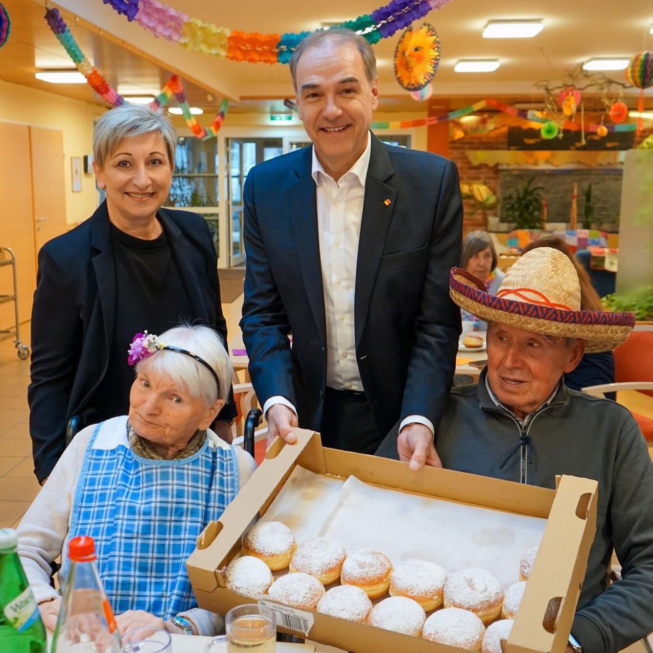 Drei Personen stehen in einem Raum mit festlicher Atmosphäre und halten eine Schachtel mit Donuts. Der Mann lächelt, während die Frau auf der linken Seite eine Stirnband trägt. Der ältere Mann rechts trägt einen Sombrero. Eine grüne Flasche und ein Glas mit Flüssigkeit stehen auf dem Tisch.