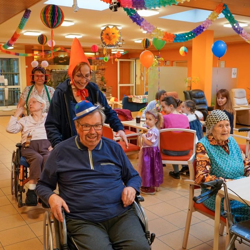 Eine Gruppe älterer Menschen und Kinder feiert eine Party in einem farbenfrohen Raum mit Luftballons und Dekorationen.