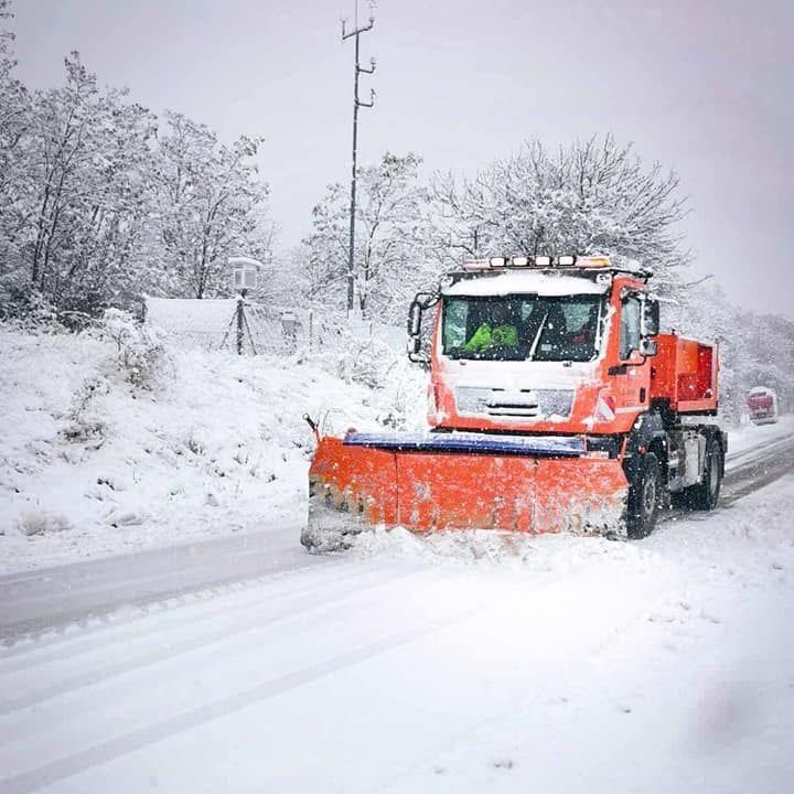 Ein orangefarbener Schneepflug räumt Schnee auf einer Straße, mit Bäumen und einem Versorgungsmast im Hintergrund.