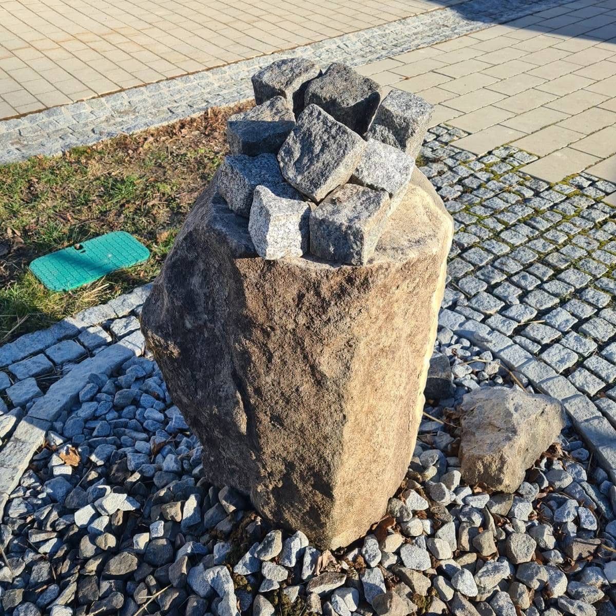 Ein großer Stein steht auf einem Kiesweg mit kleineren Steinen darauf und darum herum, in der Nähe eines grünen Teppichs.