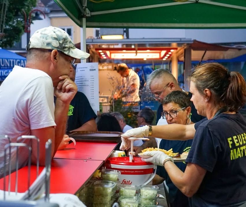 Eine Gruppe von Menschen versammelt sich um einen Imbissstand, an dem zwei Frauen in schwarzen T-Shirts Essen servieren. Der Stand hat einen roten Tresen und ein grünes Dach.