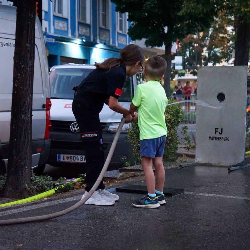 Eine Frau in schwarzer Uniform sprüht Wasser aus einem Schlauch auf einen Jungen in einem neongrünen T-Shirt und blauen Shorts.