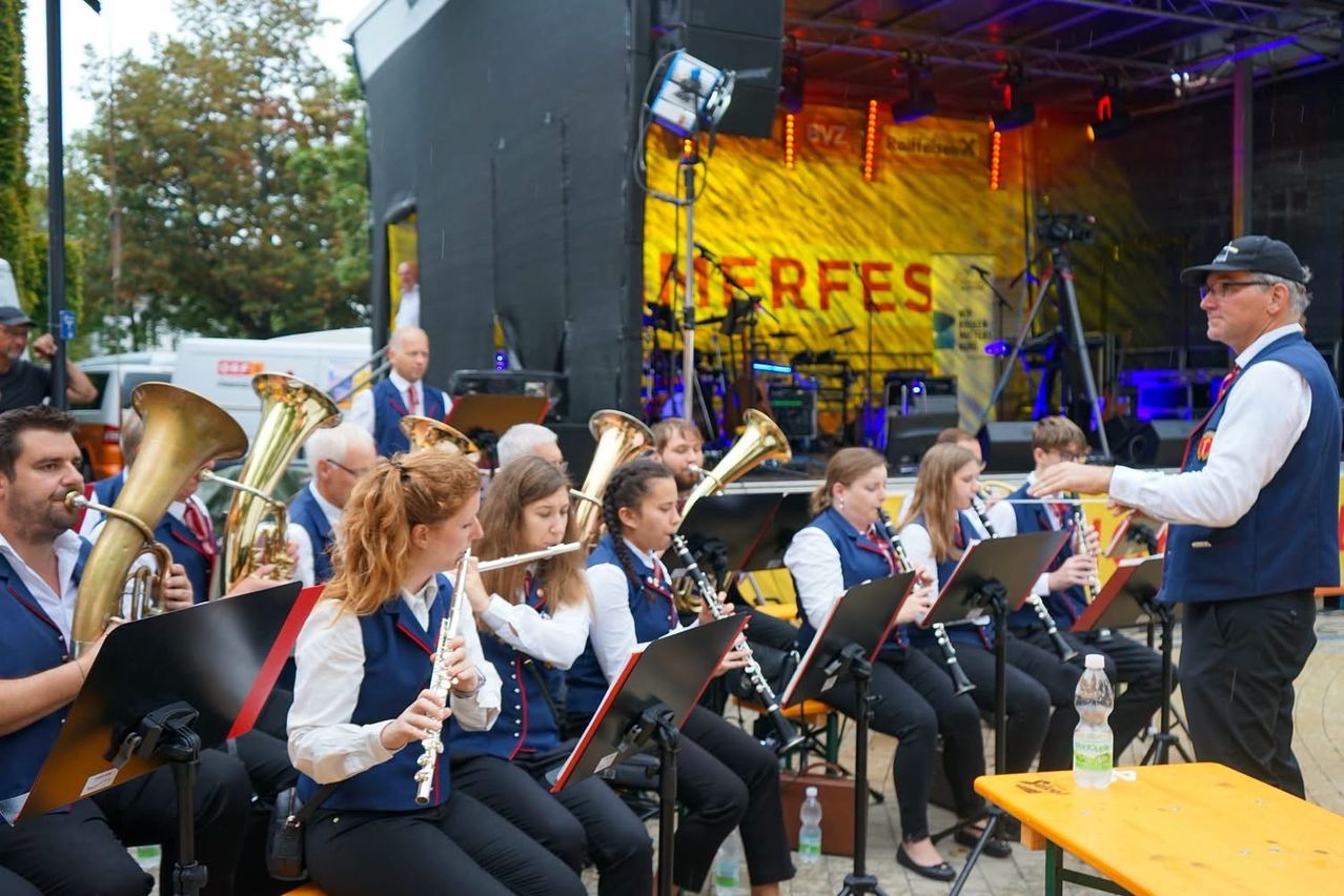 Eine Gruppe von Musikern, darunter junge Mädchen, spielt auf der Bühne mit verschiedenen Instrumenten wie Flöten und Tubas. Die Bühne ist gut beleuchtet mit einem gelben Hintergrund und einem Banner.