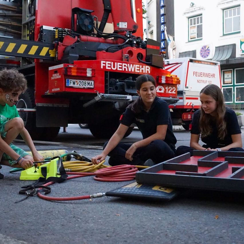 Drei Kinder sitzen auf der Straße, eines hält ein Werkzeug und zwei andere schauen auf ein Labyrinthbrett. Ein Feuerwehrwagen ist hinter ihnen geparkt.