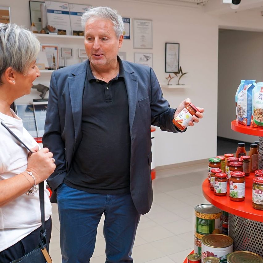 Ein Mann in einem blauen Anzug hält eine Flasche Sauce und spricht mit einer Frau in einem weißen Shirt in einem Geschäft mit Regalen voller Konserven im Hintergrund.