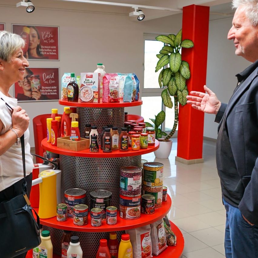 Eine Frau mit grauem Haar steht neben einem roten Regal, das mit verschiedenen Lebensmitteln gefüllt ist, während ein Mann in einem blauen Mantel zu ihr spricht. Hinter ihnen befindet sich eine Wand mit Postern und einem Topf mit Pflanzen.