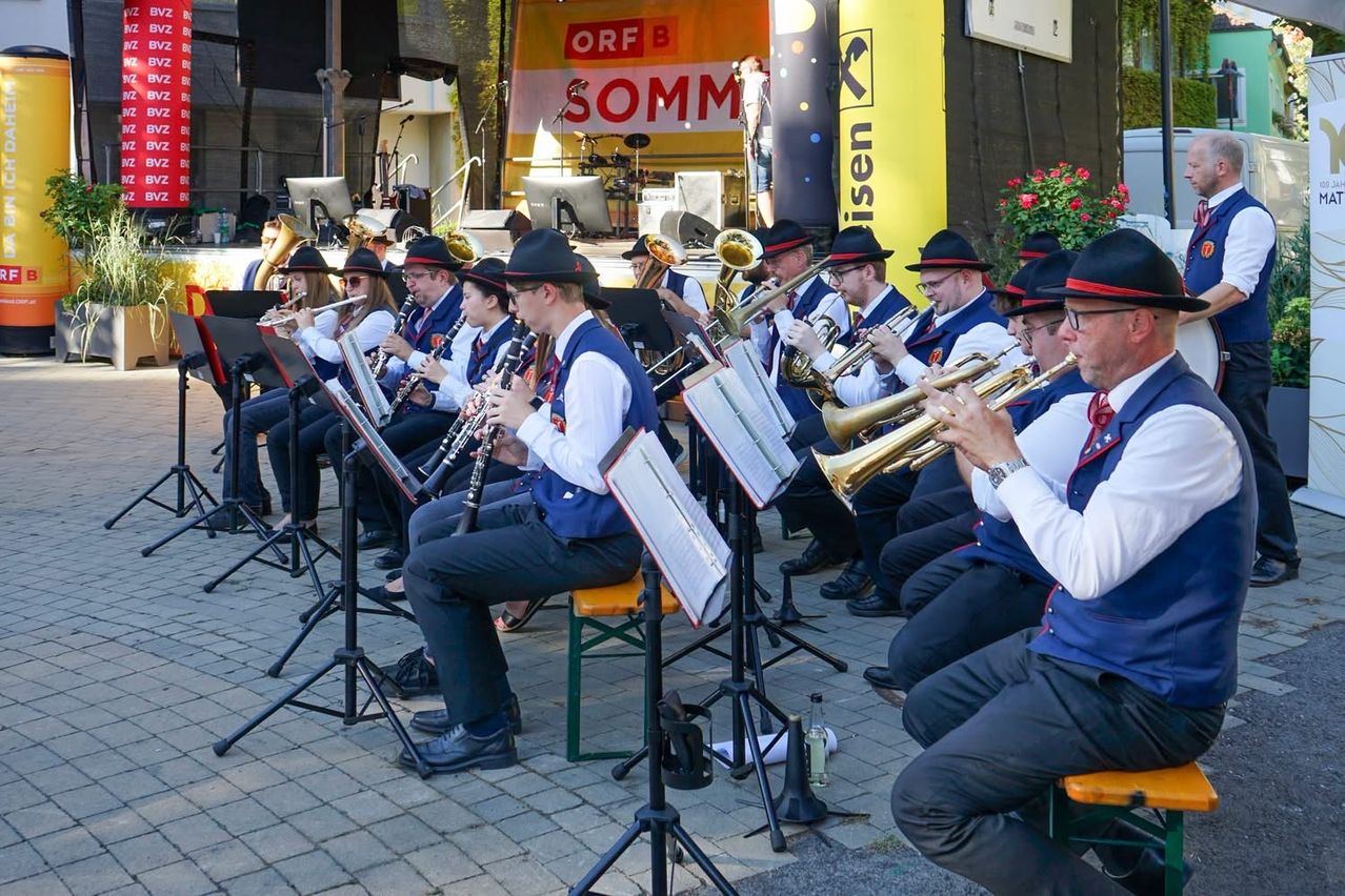 Eine Blaskapelle spielt auf einer Bühne mit einem Hintergrund, der den Text 'ORF B SOMM' zeigt. Die Musiker sind in traditionellen Uniformen gekleidet und spielen verschiedene Instrumente.
