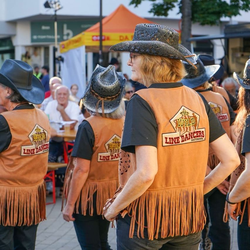 Eine Gruppe von Menschen in Cowboyhüten und Lederwesten, beschriftet mit 'Line Dancers', gehen in einer Reihe. Sie befinden sich in einem Außenbereich mit Zelten und Gebäuden im Hintergrund.