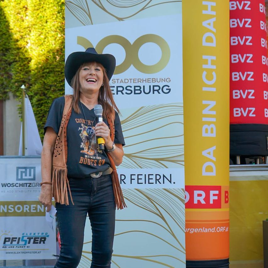Eine Frau in einem Cowboyhut und einem gefütterten Weste steht vor einem Banner mit dem Text 'Stadterhebung Fersbrug' und spricht in ein Mikrofon.
