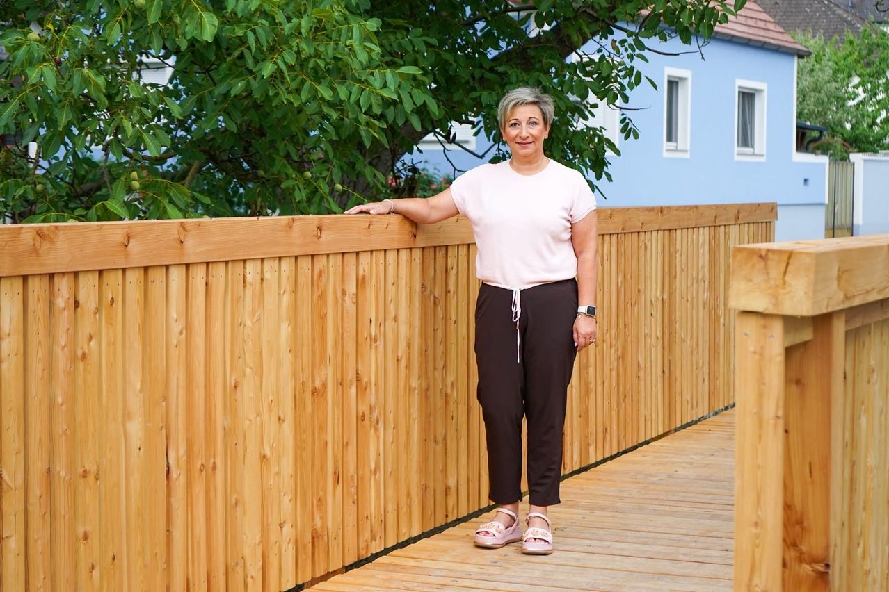 Eine Frau mit grauen Haaren steht auf einer Holzterrasse, lächelt und lehnt sich an einen Holzzaun. Sie trägt ein rosa Oberteil, braune Hosen und rosa Sandalen. Hinter ihr befindet sich ein blaues Haus mit weißen Fenstern und einem Baum.