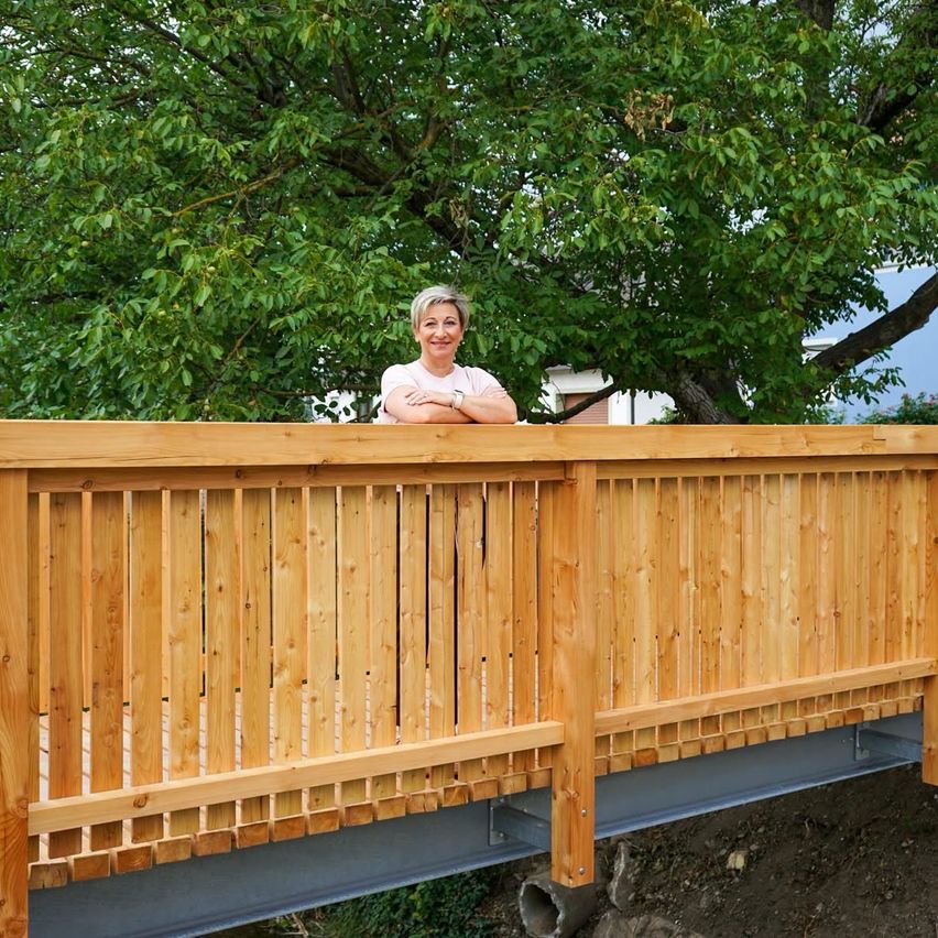 Eine Frau steht mit verschränkten Armen lächelnd auf einer Holzterrasse, mit einem großen Baum im Hintergrund.