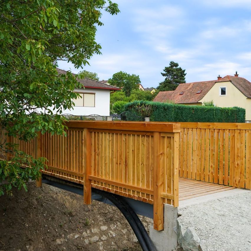 Eine Holzbrücke mit einem Holzgeländer und einem Betonfundament ist über einem Erdweg errichtet. Dahinter befinden sich Häuser mit braunen Dächern und einer Hecke davor.