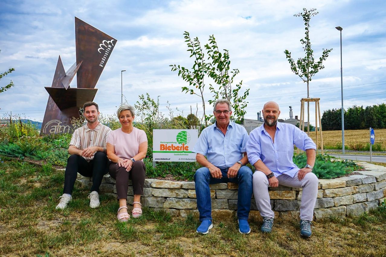 Vier Personen sitzen auf einer Steinmauer vor einem Garten und lächeln für ein Foto. Dahinter steht ein Schild mit dem Text 'Bieberle' und eine Metallskulptur. Bäume und Pflanzen sind im Hintergrund.
