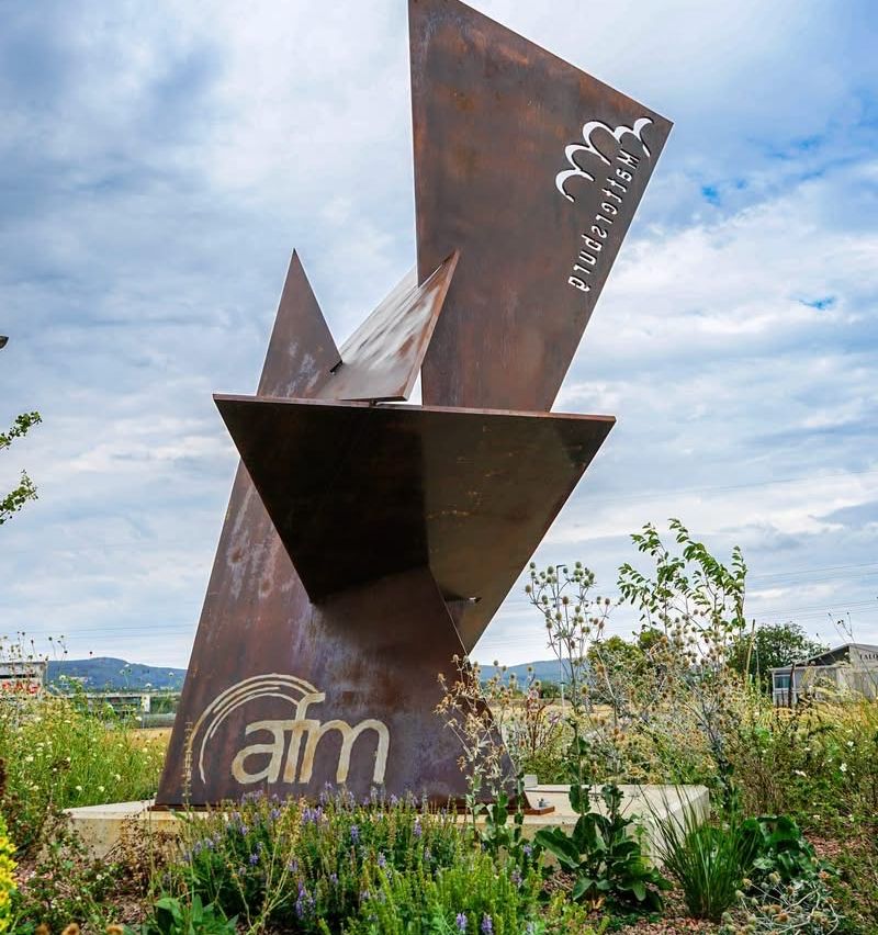 Eine große, abstrakte Metallskulptur mit dem Wort 'atm' im Vordergrund. Die Skulptur steht in einem Feld mit verschiedenen Pflanzen und einem bewölkten Himmel im Hintergrund.