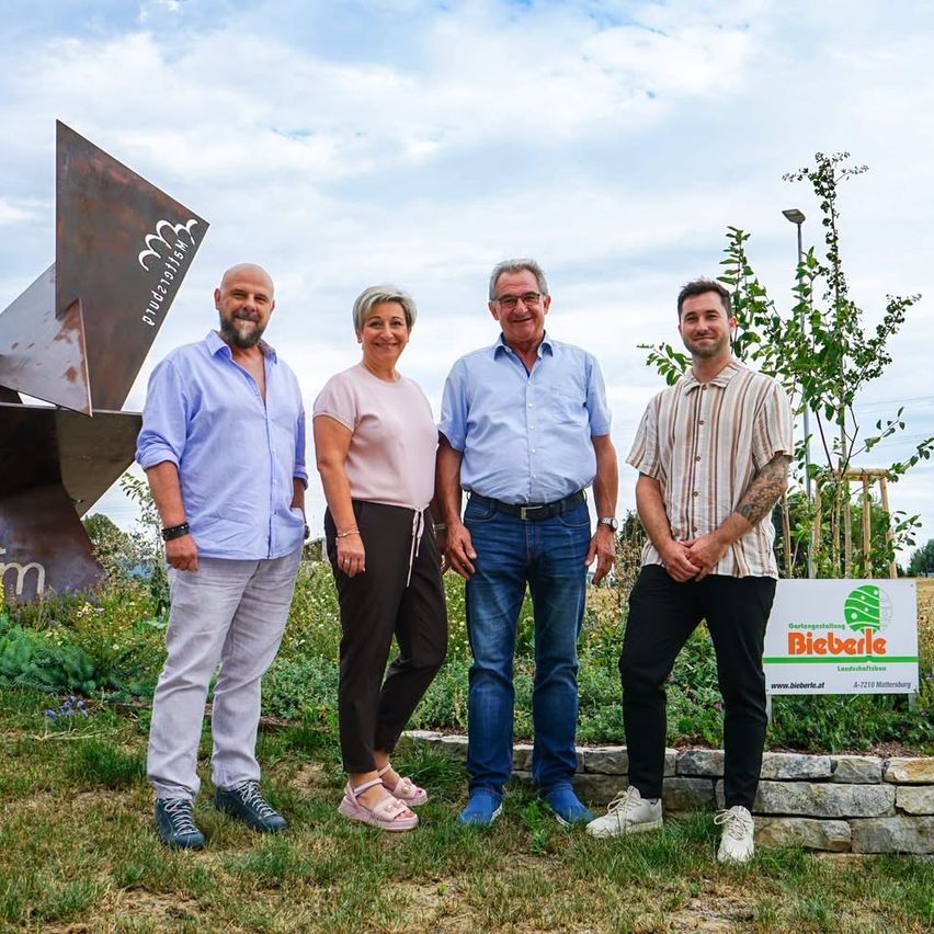 Vier Personen stehen vor einem Garten mit einem Schild, auf dem 'Bieberle' steht. Sie lächeln und scheinen für ein Foto zu posieren. Dahinter steht eine große Metallskulptur.