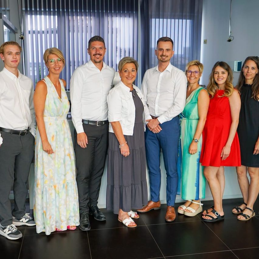 Eine Gruppe von Menschen, wahrscheinlich Kollegen, steht zusammen in einem Raum und lächelt für ein Foto. Sie sind in formeller und halbformeller Kleidung gekleidet.