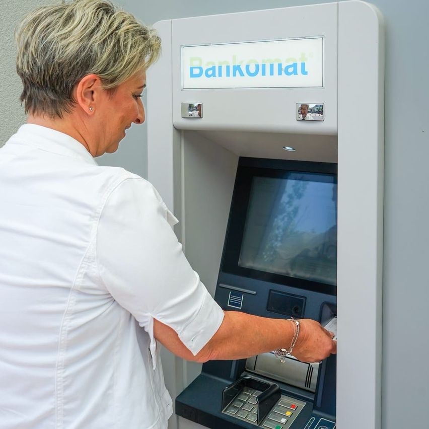Eine Frau in einem weißen Hemd benutzt einen Bankomat, der 'Bankomat' beschriftet ist, und hält eine Karte in der Hand.