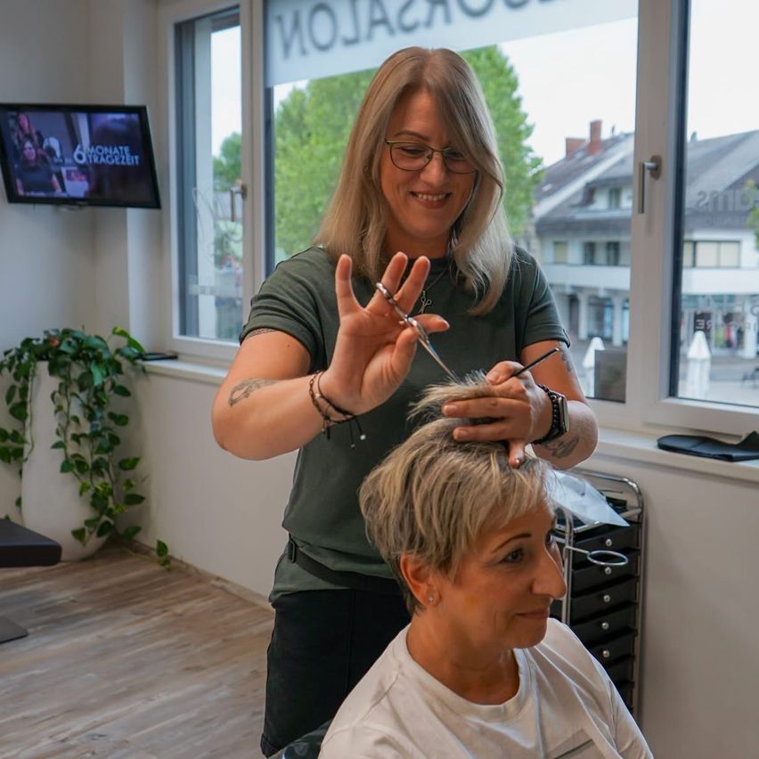 Eine Frau mit Brille und grünem Shirt schneidet die Haare einer anderen Frau, die in einem Salon sitzt. Im Hintergrund sind eine Pflanze und ein Fernseher zu sehen.