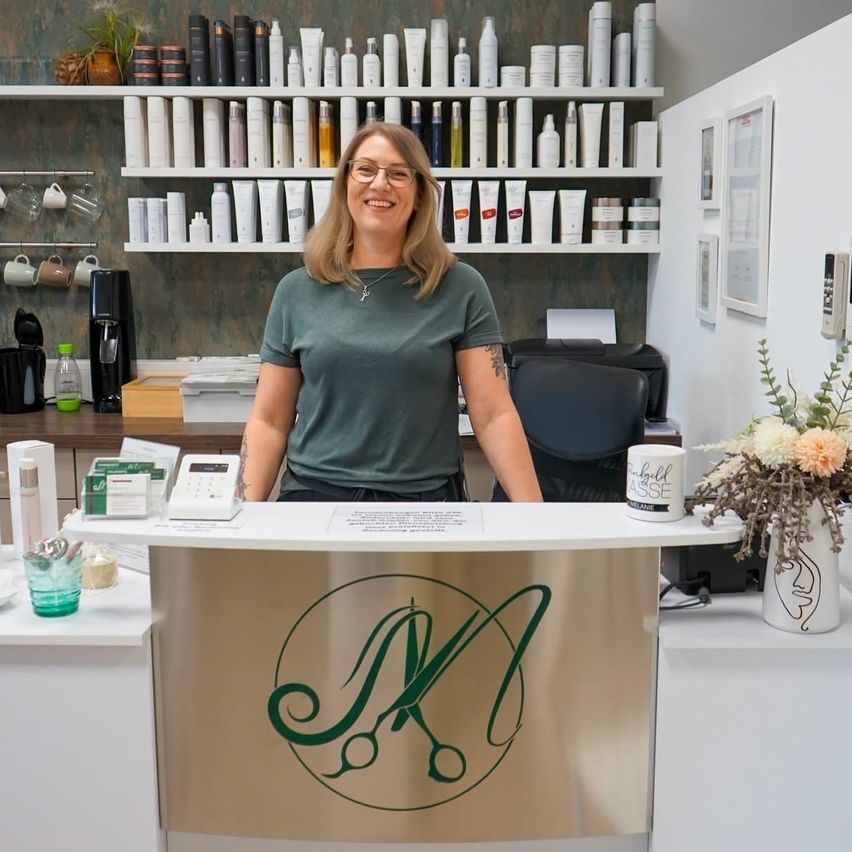Eine Frau steht lächelnd hinter einer Rezeptionstheke in einem Salon. Die Theke hat ein Logo und eine Vase mit Blumen. Hinter ihr befinden sich Regale mit verschiedenen Schönheitsprodukten.
