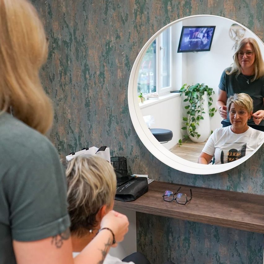 Eine Frau mit blonden Haaren lässt sich in einem Salon die Haare machen. Sie sitzt vor einem Spiegel mit dem Reflex einer anderen Frau und einer Pflanze.