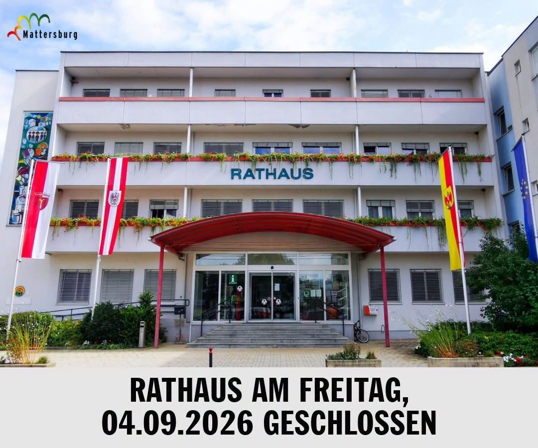 The front of a white building with the word Rathaus on top, with flags on the side and a red awning over the entrance.