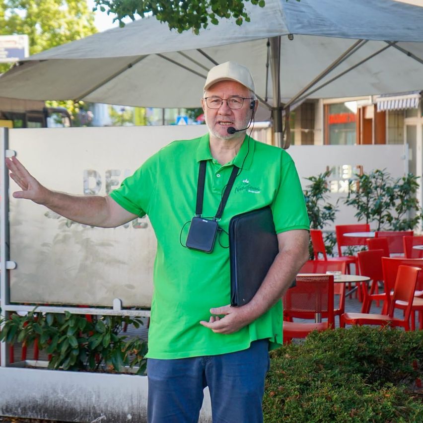 A man in a green shirt and blue jeans stands outside, holding a folder and wearing a headset. He is gesturing with his hand and appears to be speaking. Behind him is a large umbrella, a wall, and a table with red chairs.