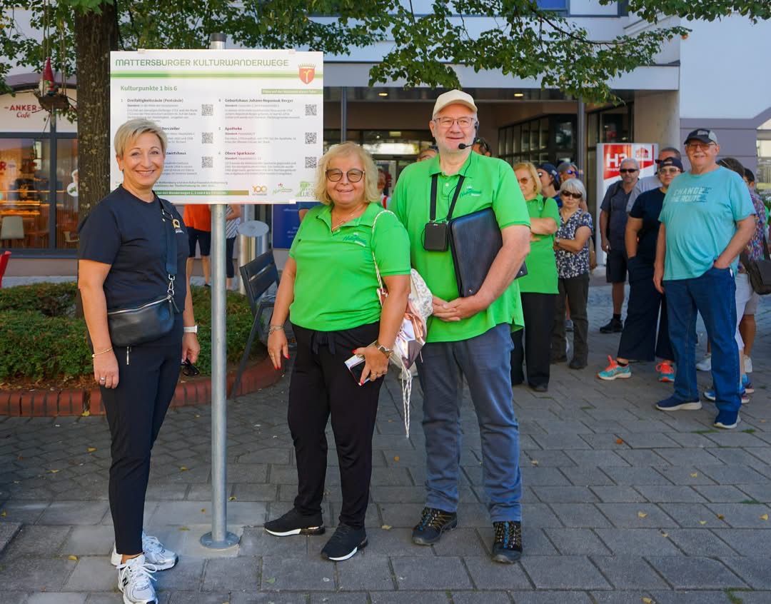 Three people in green shirts stand in front of a sign on a sidewalk, with a crowd of people in the background.