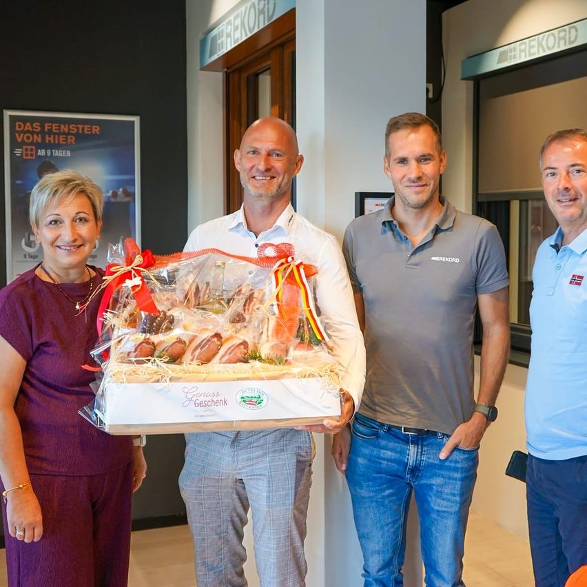 Four people stand together, smiling. A man in the center holds a gift basket with wrapped food items. Behind them is a sign that reads 'Das Fenster von Hier'.