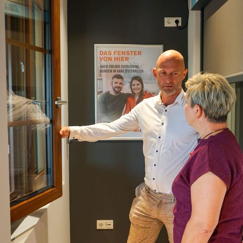 A man and a woman are standing in front of a glass door, with the man pointing at the door. Behind them is a wall with a poster advertising a company.