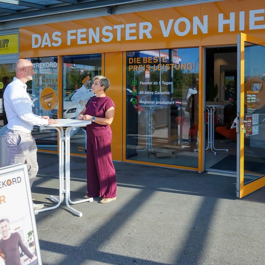 Two people stand outside a store with a sign that reads 'Das Fenster von hier'. The man wears a white shirt and the woman a purple jumpsuit. They are near a table with a sign on it.