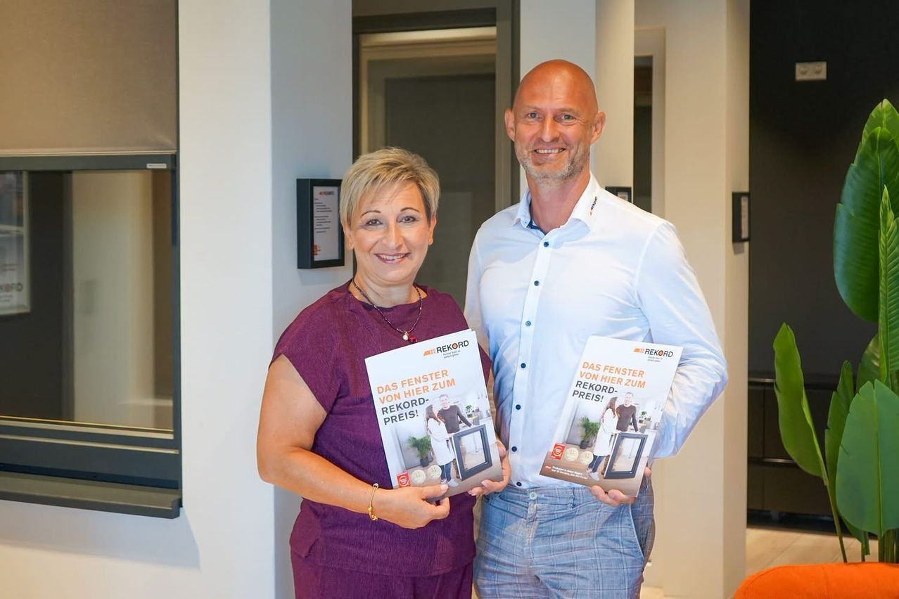A man and a woman stand together, smiling, holding up brochures titled 'Das Fenster von hier zum Rekord-Preis!' The woman wears a purple dress and a necklace, while the man wears a light blue shirt and jeans. Behind them is a white wall with a framed picture.