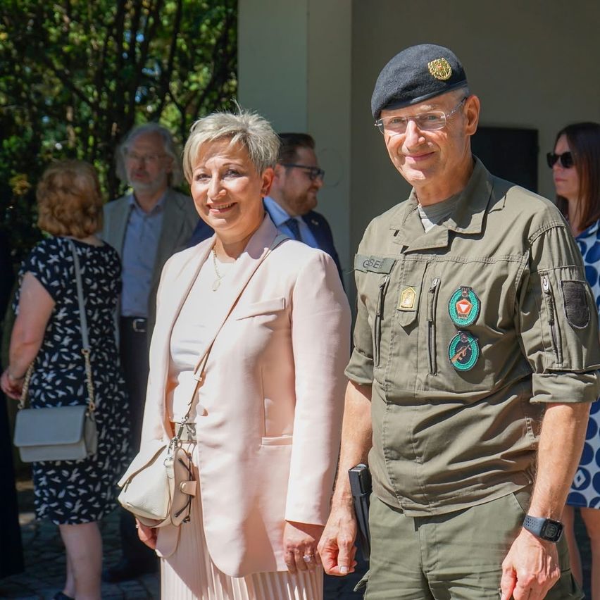 Ein Mann in Uniform und eine Frau in einem rosa Anzug stehen nebeneinander. Sie lächeln und schauen in die Kamera.