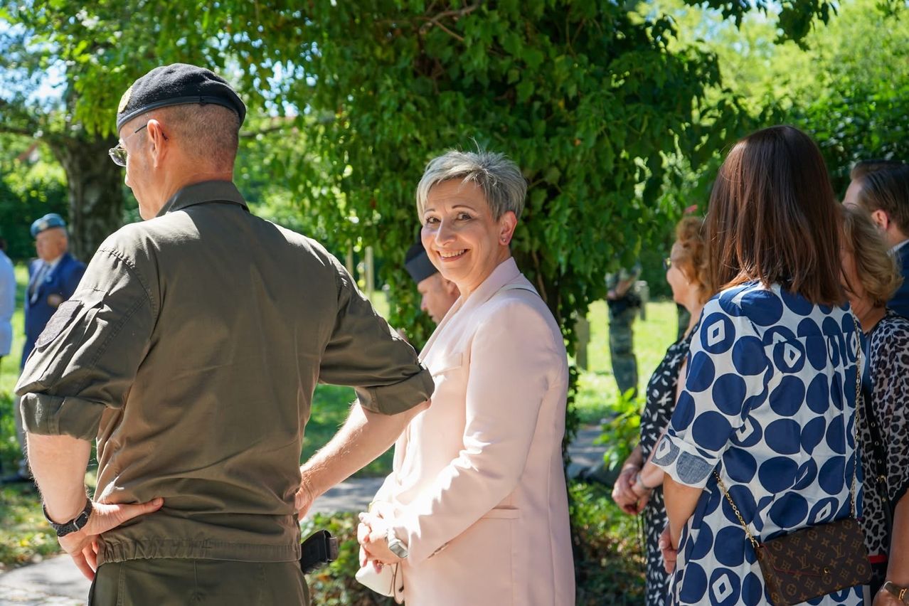 Eine lächelnde Frau in einer weißen Jacke steht neben einem Mann in Uniform, sie sind von anderen umgeben, ein Baum und ein Pfad im Hintergrund.