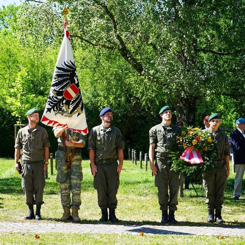 Fünf uniformierte Männer stehen in einer Reihe auf einem Weg. Sie halten eine Flagge und einen Kranz. Hinter ihnen steht ein Mann in einem Anzug. Bäume sind hinter den Männern.