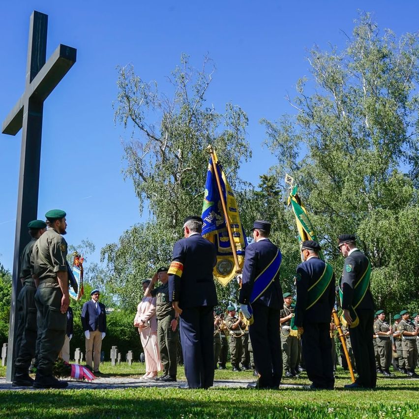 Eine Gruppe uniformierter Personen steht um ein Kreuz herum, einige mit Fahnen, bei einer Gedenkfeier. Eine Band spielt im Hintergrund.