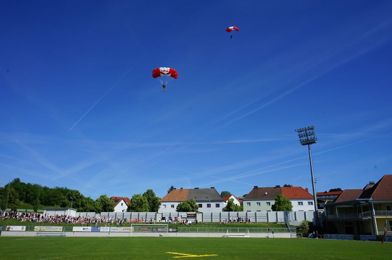Zwei Personen paragliden am Himmel über einem Fußballfeld, während eine Menge hinter einer Mauer zuschaut. Gebäude und Bäume sind im Hintergrund.