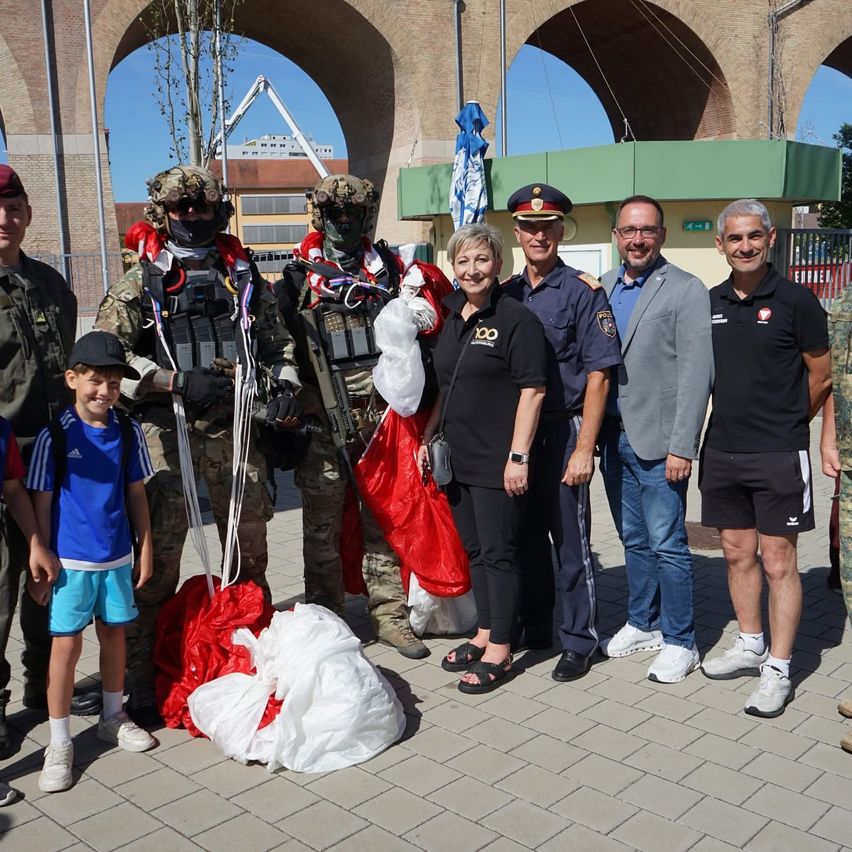 Eine Gruppe von Menschen, darunter einige in Militärausrüstung, stehen lächelnd für ein Foto auf einem gepflasterten Weg. Ein Kind in einem blauen T-Shirt steht neben einem Soldaten. Hinter ihnen ist ein Gebäude mit grünen Verzierungen und einem gewölbten Eingang sichtbar.