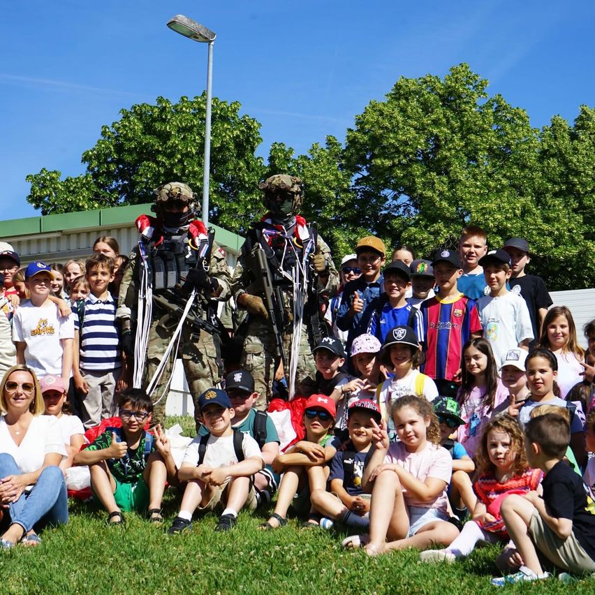 Gruppenfoto von Soldaten mit Kindern, zwei Erwachsene und mehrere Kinder sitzen auf dem Rasen, während zwei Soldaten in voller Ausrüstung hinter ihnen stehen.